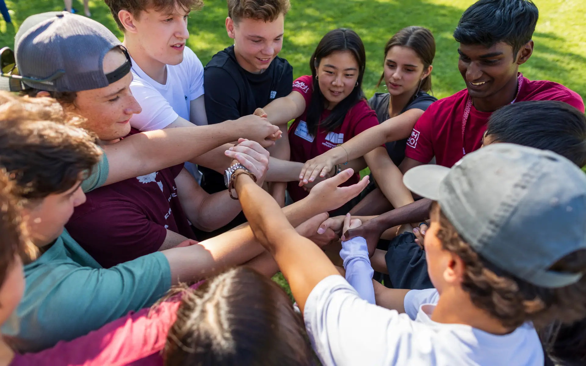 Group of diverse young people standing in a circle outdoors with their hands joined together in the center.