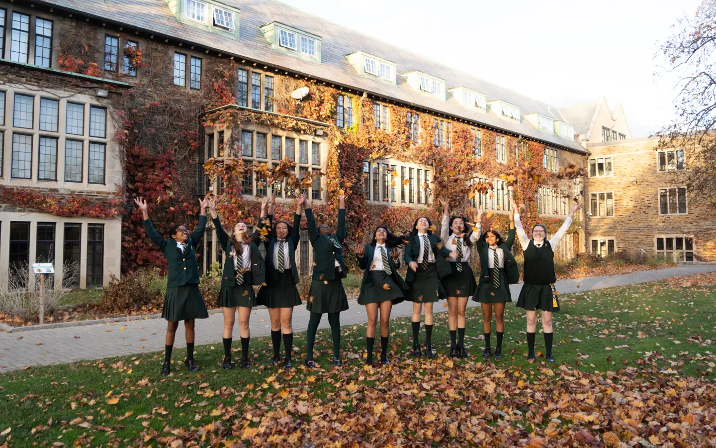 Group of students in green school uniforms joyfully tossing autumn leaves in front of a large ivy-covered school building.
