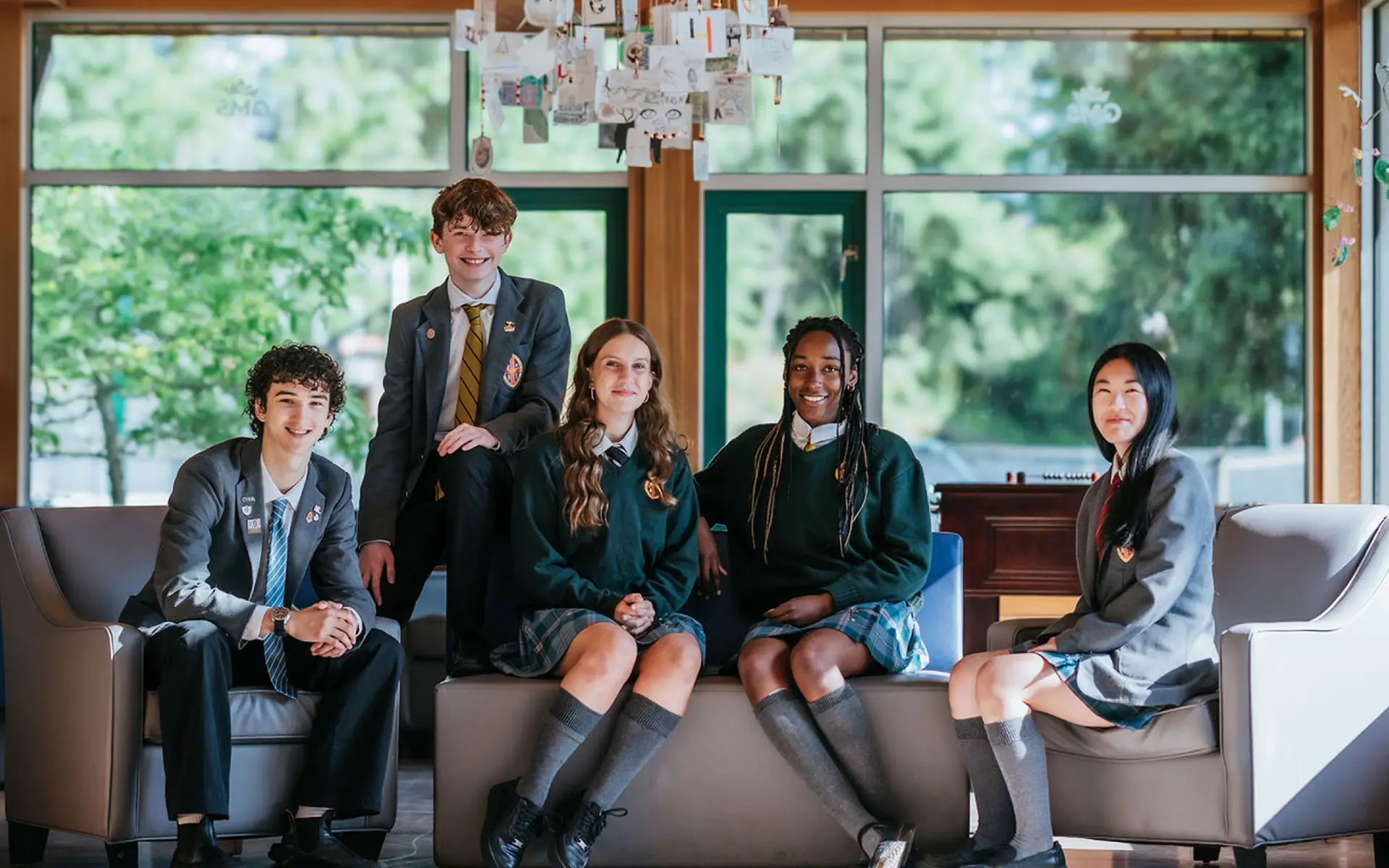 Five high school students in uniforms sitting and smiling in a bright room with large windows and greenery outside.