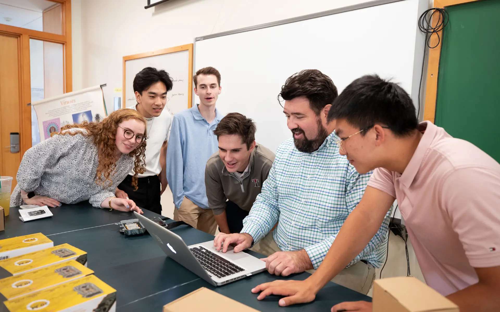 Group of six people gathered around a laptop in a classroom, engaged and smiling during a collaborative activity.