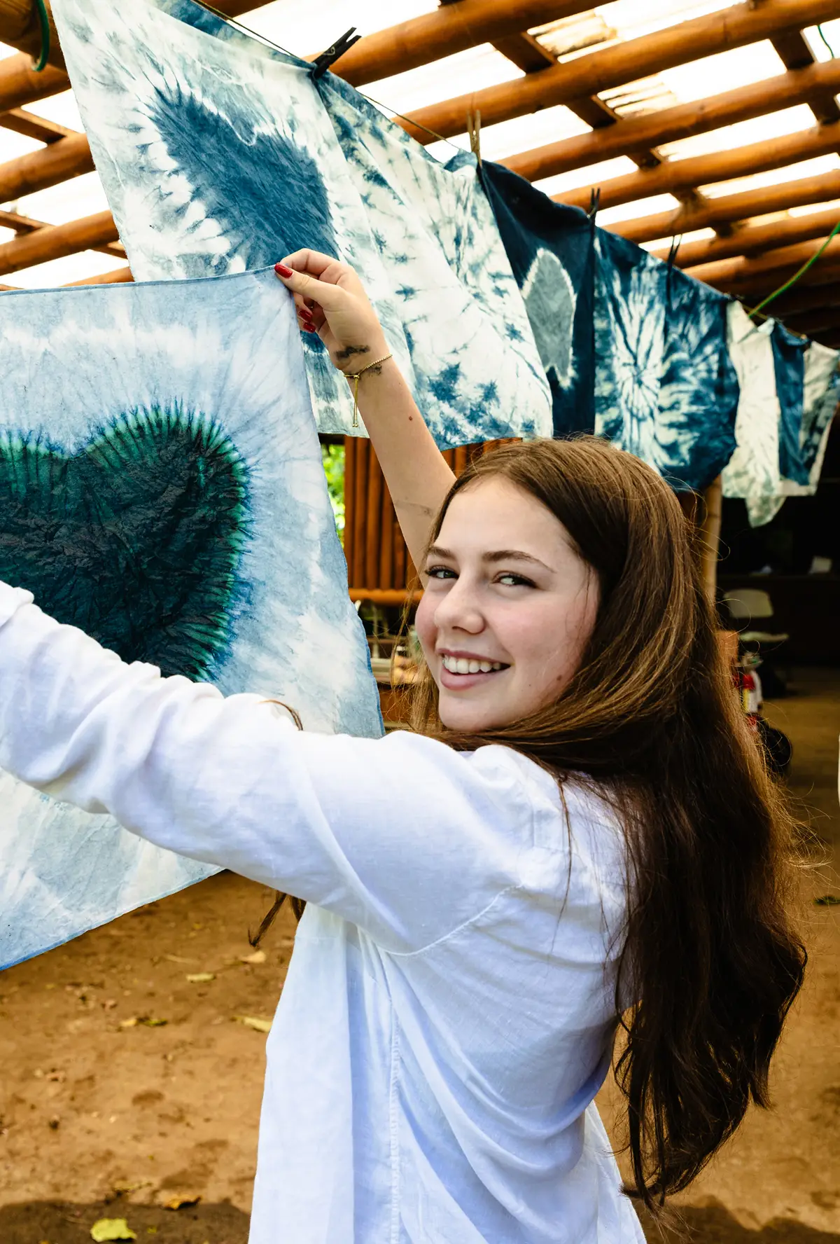 Smiling young woman hanging blue tie-dye fabric with heart patterns on a clothesline under a wooden pergola.