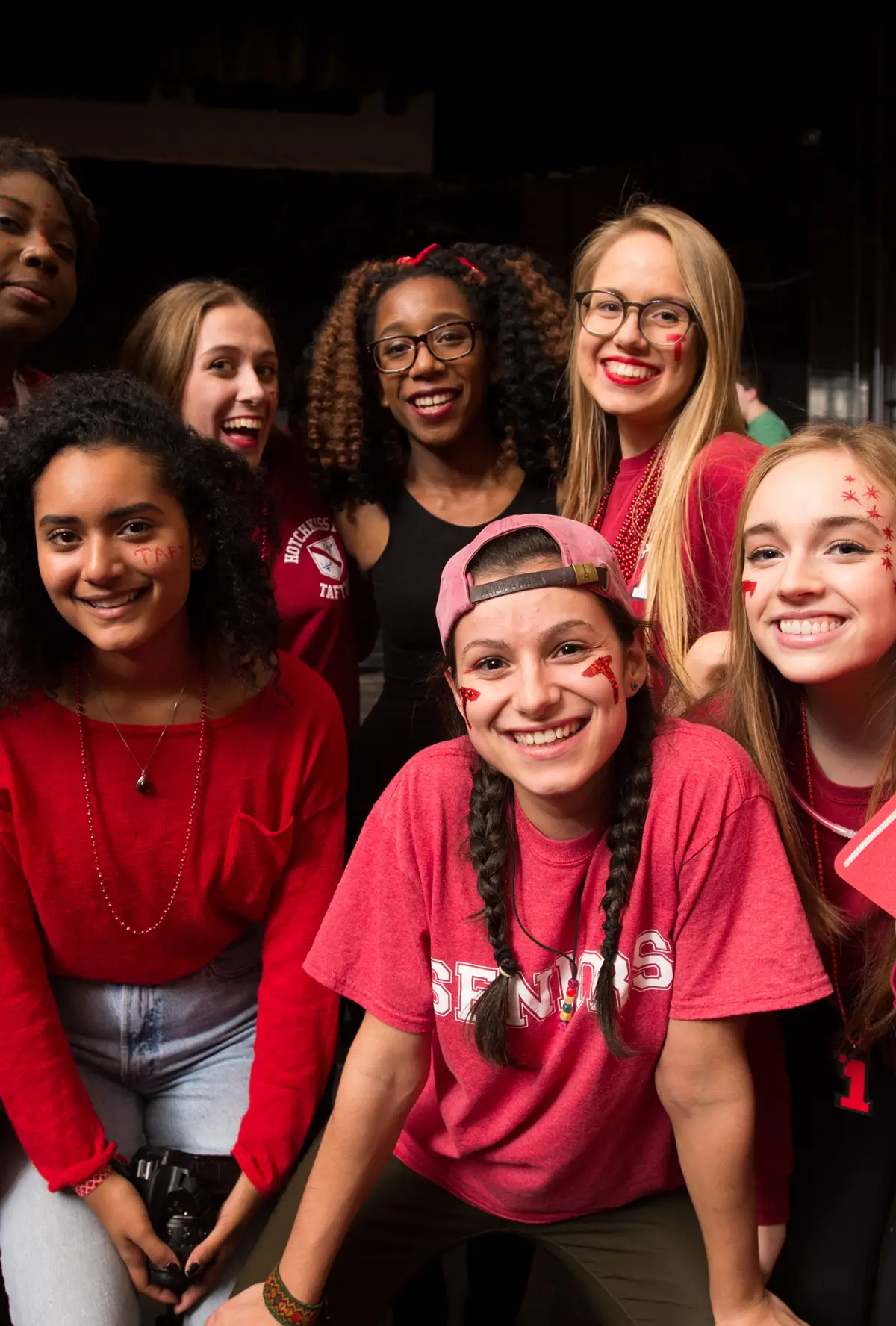 Group of smiling young women wearing red and pink, some with face paint and beads, posing together indoors.