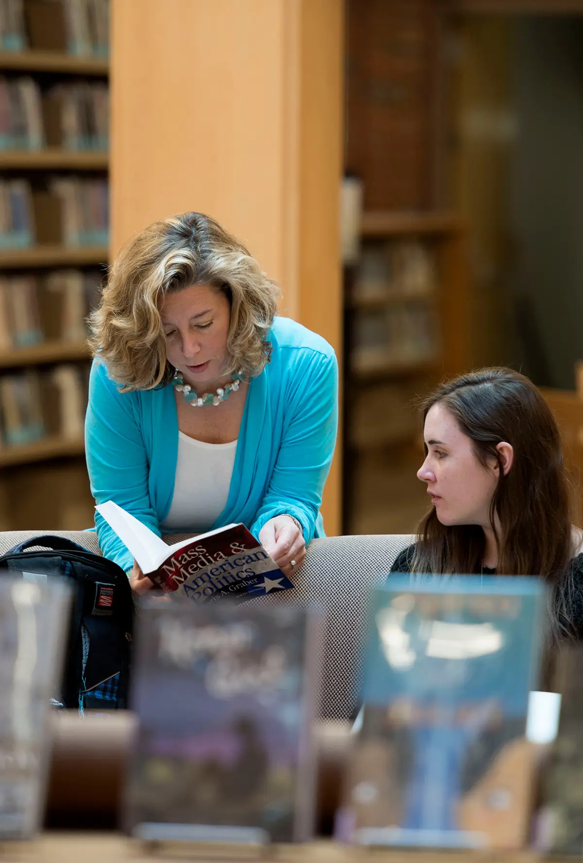 A woman in a blue cardigan reads a book titled 'Mass Media & American Politics' to a younger woman sitting nearby in a library.