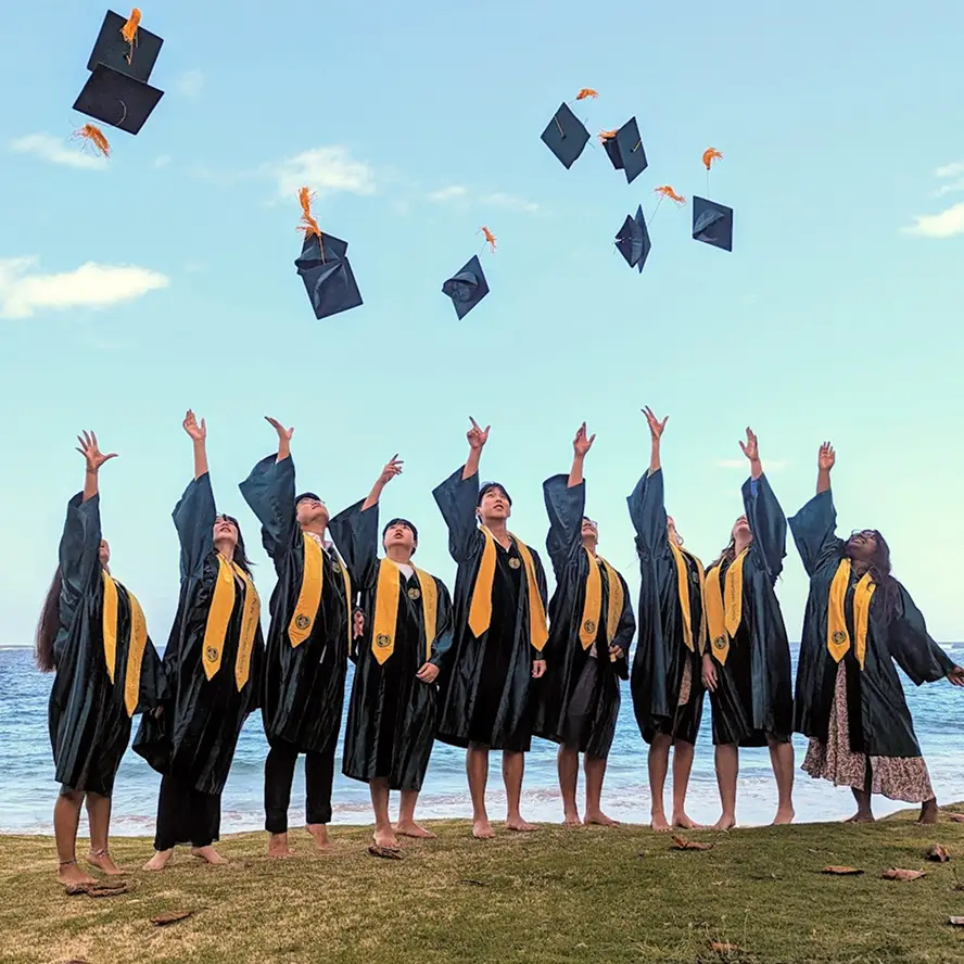 Group of graduates in black gowns and yellow stoles tossing their caps in the air by the ocean shore.