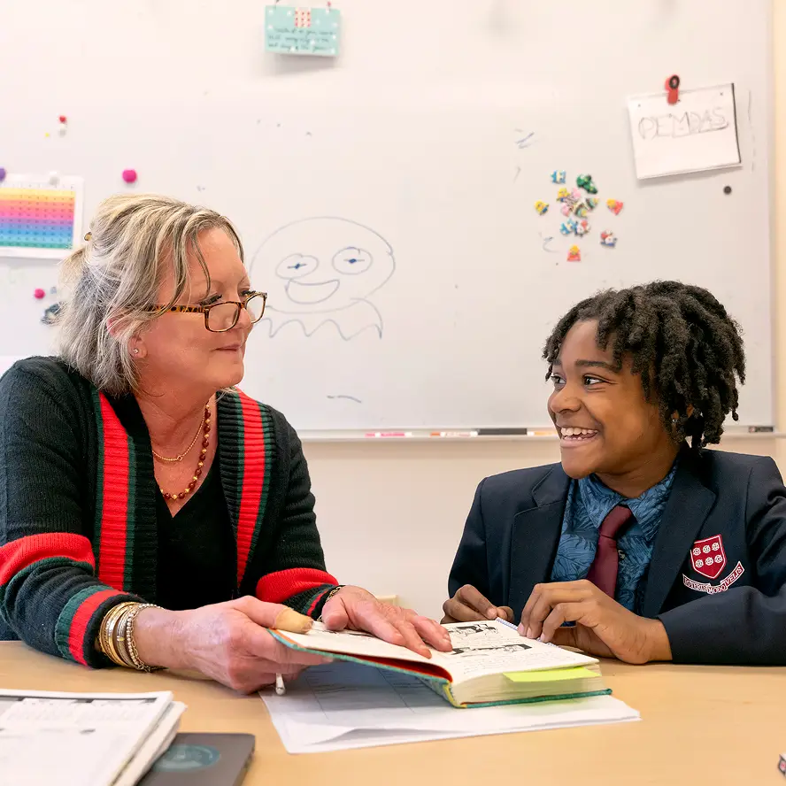 Teacher and student smiling and discussing a book at a desk with a whiteboard in the background.