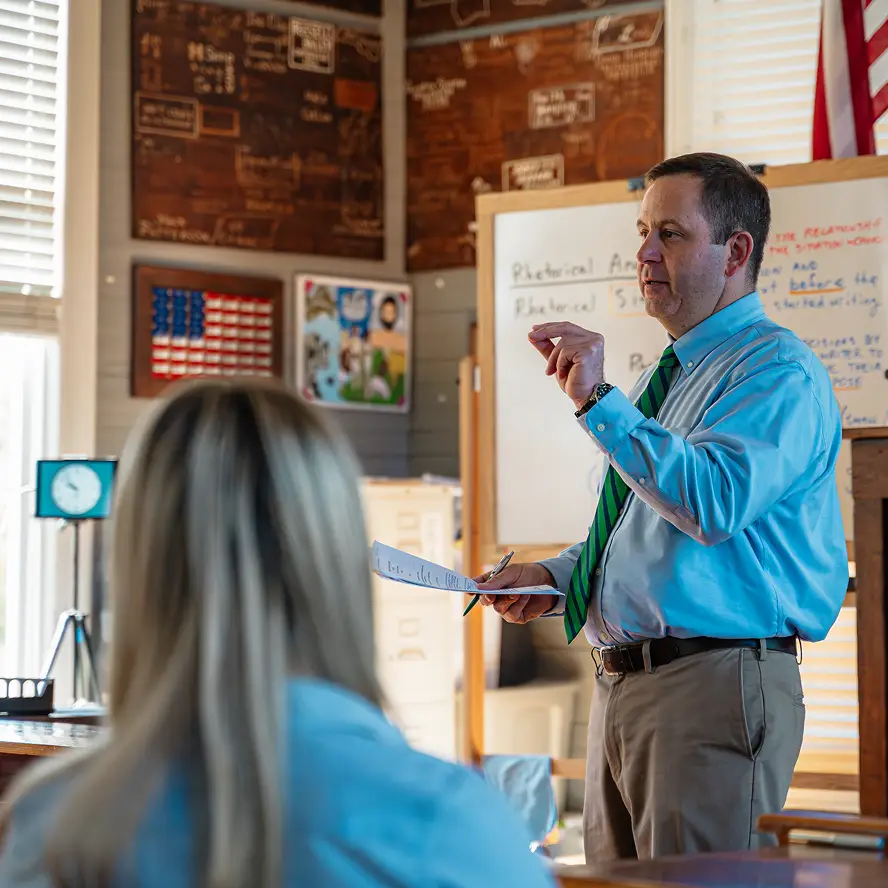 Teacher in blue shirt and green striped tie speaking in front of a whiteboard with handwritten notes in a classroom.