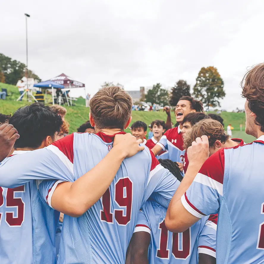 Group of young soccer players in blue and red uniforms celebrating together outdoors on a cloudy day.