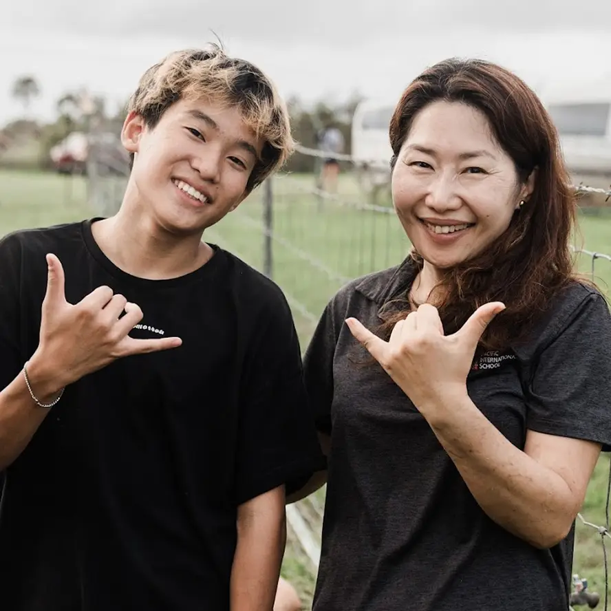 Smiling young man and woman outdoors making shaka hand gestures near a wire fence.