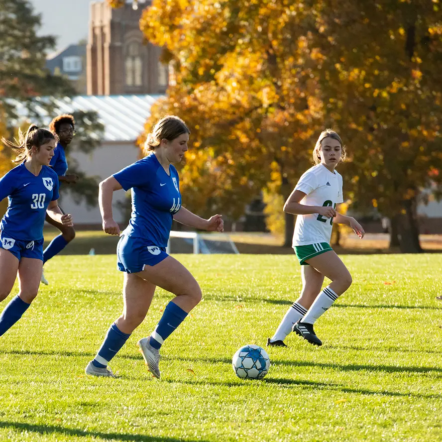 Girls' soccer players from two teams competing for the ball on a sunny field with autumn trees in the background.