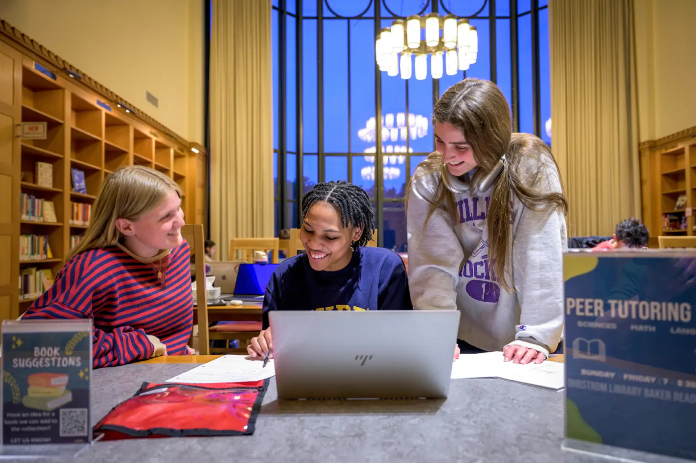 Three students studying together with a laptop and papers in a library with large windows and bookshelves.