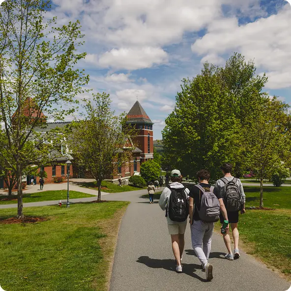 Three students walking on a paved path through a green campus with brick buildings and trees under a partly cloudy sky.