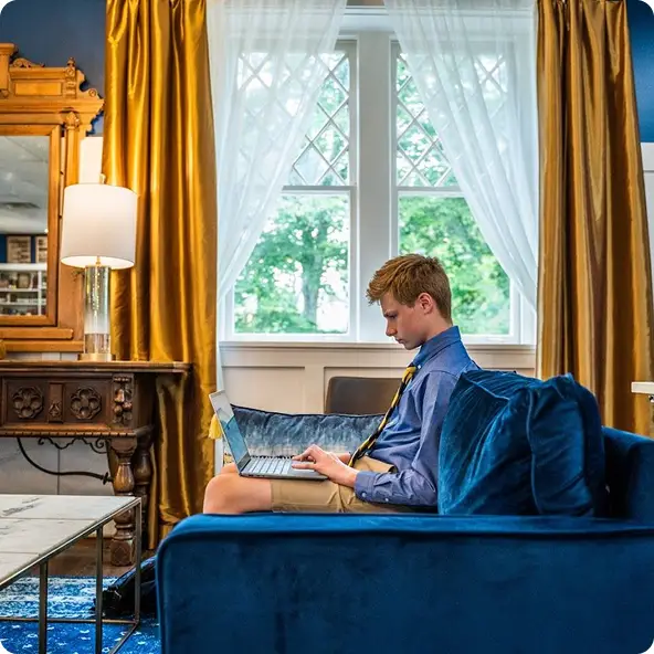 Boy in a blue shirt and tie sitting on a blue couch using a laptop in a room with gold curtains and a window with white sheer curtains.