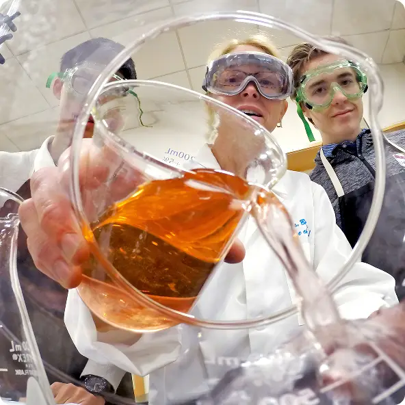 Three people wearing safety goggles and lab coats conducting a chemistry experiment with orange liquid in a beaker.