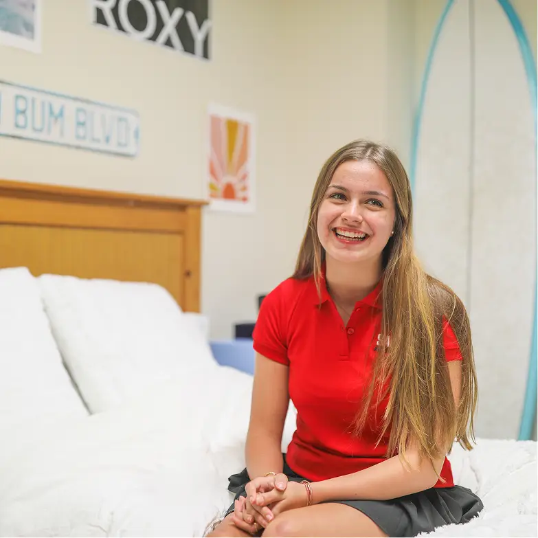 Smiling young woman with long blonde hair wearing a red polo shirt sitting on a white bed in a bedroom.