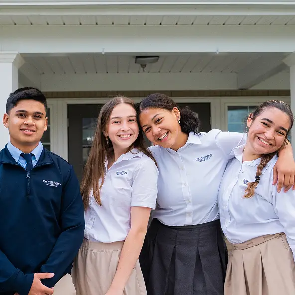 Four teenagers in school uniforms smiling and standing together with their arms around each other outside a building.