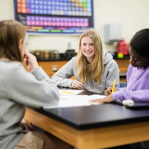 Three students sitting around a table in a classroom, with one girl smiling at the camera and a periodic table visible in the background.