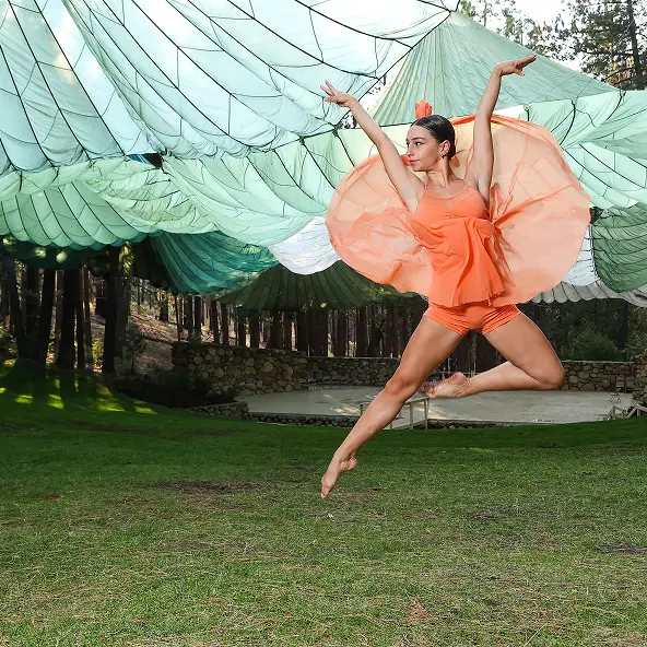 Female dancer in an orange outfit performing a mid-air leap on green grass with large parachutes suspended overhead and trees in the background.