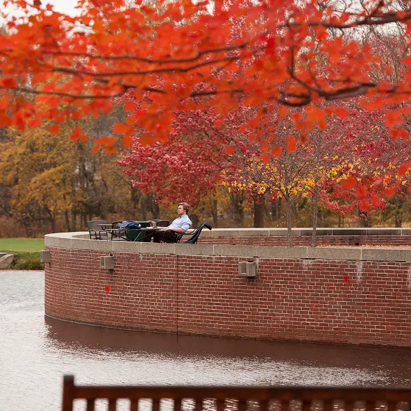 Person sitting on a chair on a curved brick platform by a pond, surrounded by vibrant red and orange autumn trees.