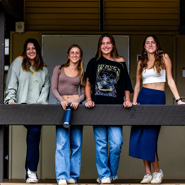 Four young women standing behind a wooden railing, smiling and casually dressed in jeans and tops.