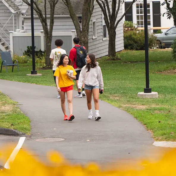 Two young women walking and talking on a paved path through a green lawn with residential buildings and trees in the background.