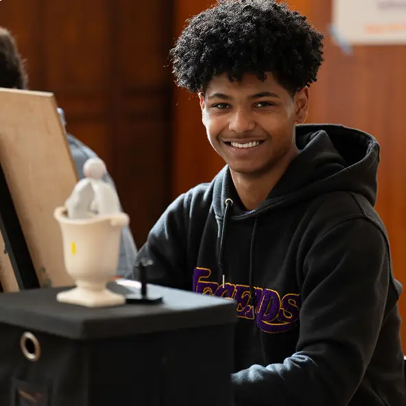 Smiling young man with curly hair wearing a black hoodie sitting at a table with an art easel and a small sculpture.