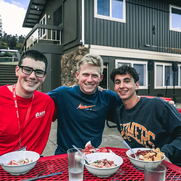 Three young men sitting at a red picnic table outdoors, smiling with bowls of food and glasses of water in front of them.