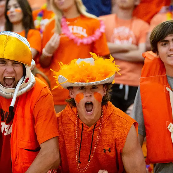 Three enthusiastic young sports fans dressed in orange, with one wearing a bright yellow and orange helmet, another in an orange feathered hat, both shouting excitedly.