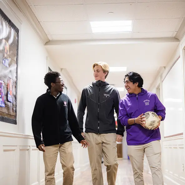 Three teenage boys walking and chatting in a school hallway, one holding a worn soccer ball.