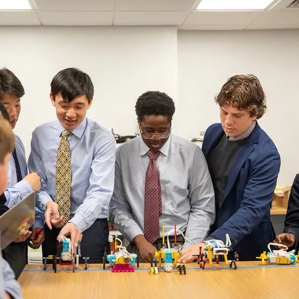 Four teenage boys in dress shirts and ties working together on colorful robotic models displayed on a table in a classroom.