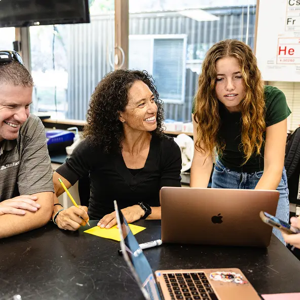 Three people gathered around a table with laptops, one woman smiling and taking notes, another woman standing and looking at the screen, and a man sitting and smiling.