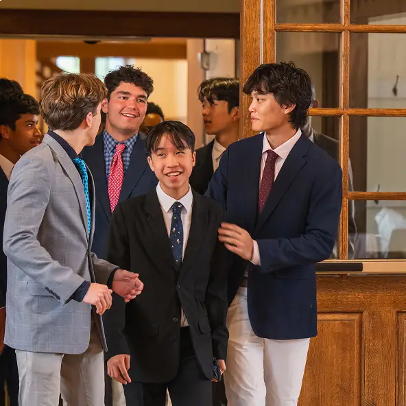Group of six teenage boys in suits and ties standing and smiling near a wooden door indoors.