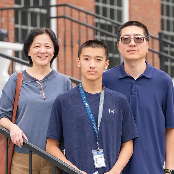 Teenage boy wearing a navy blue shirt and school ID stands between a smiling woman and a man, all posing on outdoor stairs with brick building background.