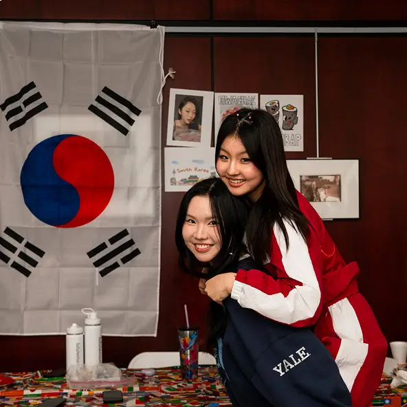 Two young women smiling and posing with one giving the other a piggyback, with a South Korean flag and cultural decorations in the background.