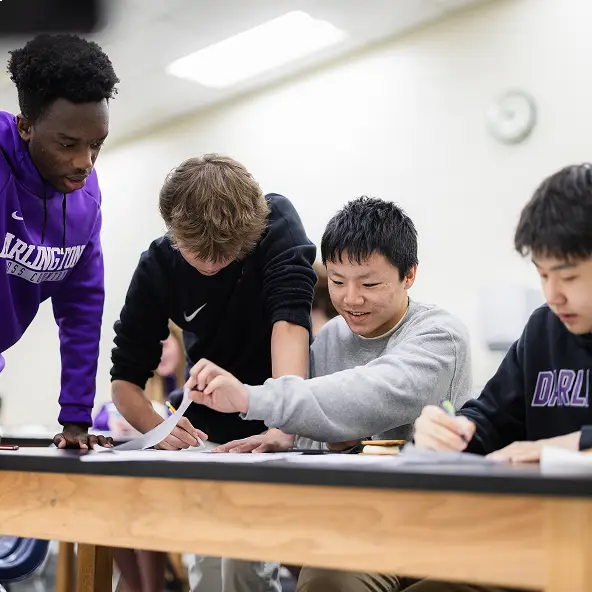 Four students collaborate at a table in a classroom, reviewing papers and writing notes together.