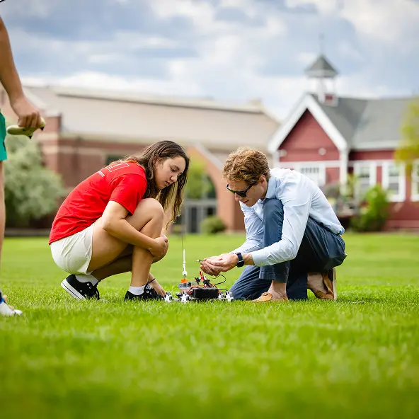 Two people crouching on grass outdoors, closely examining and adjusting a small robotic device, with buildings in the background.