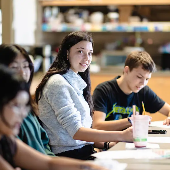 Young woman in a gray sweater sitting at a table with two other students, one writing and one blurred, in a classroom setting.