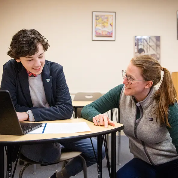 A student and a teacher smiling and interacting at a table with a laptop and papers in a classroom.