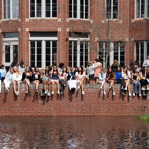 Group of young people sitting and chatting on a brick ledge by a pond in front of a large brick building with tall windows.