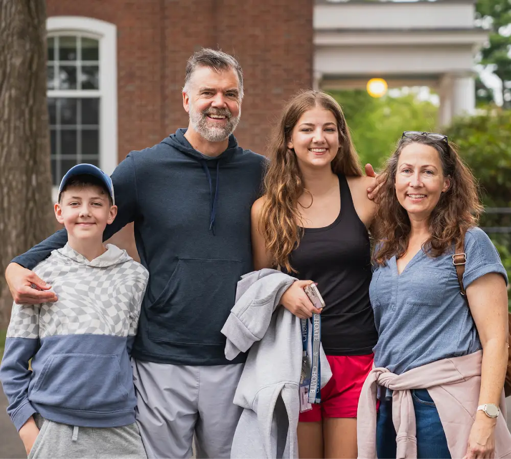 Smiling family of four outdoors, with a teenage girl, a younger boy, and two adults standing closely in casual clothing.