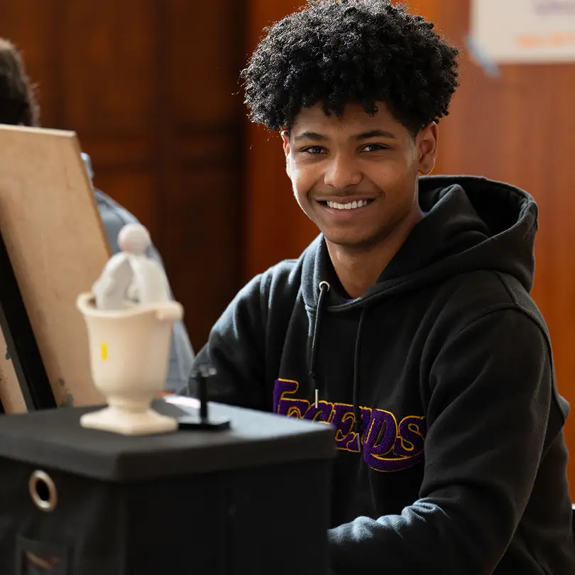 Smiling young man with curly hair wearing a black hoodie sitting at a table with art supplies.