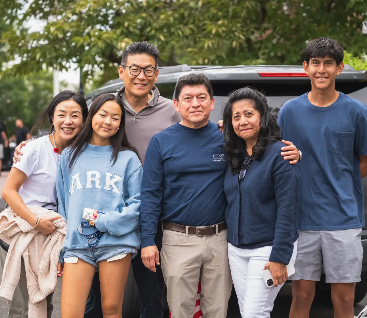Group of six people standing outside near a vehicle, smiling and posing for the photo with trees in the background.