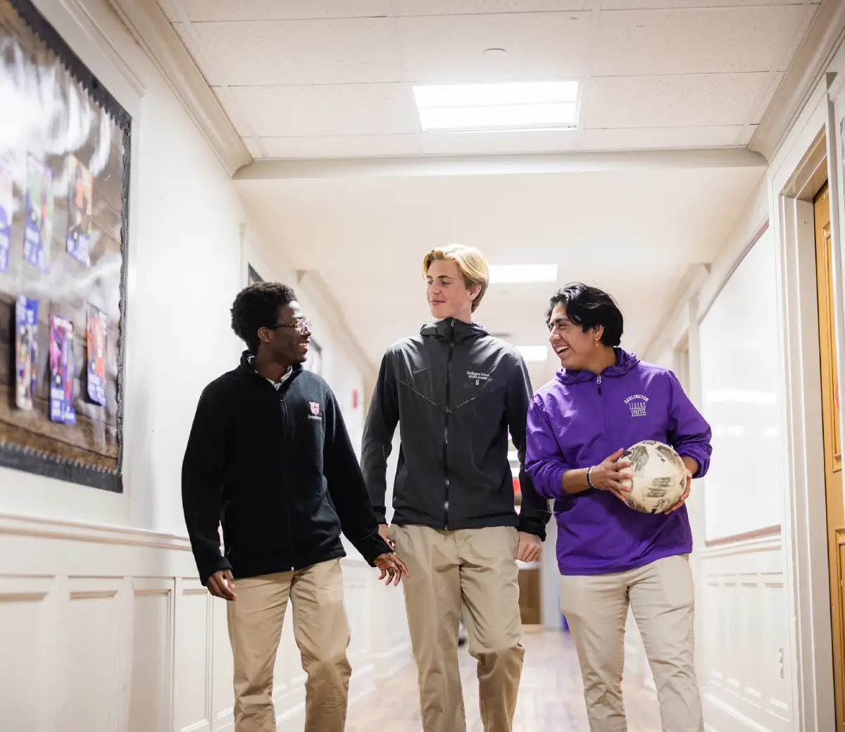 Three high school students walking in a hallway, smiling and talking, one holding a soccer ball.