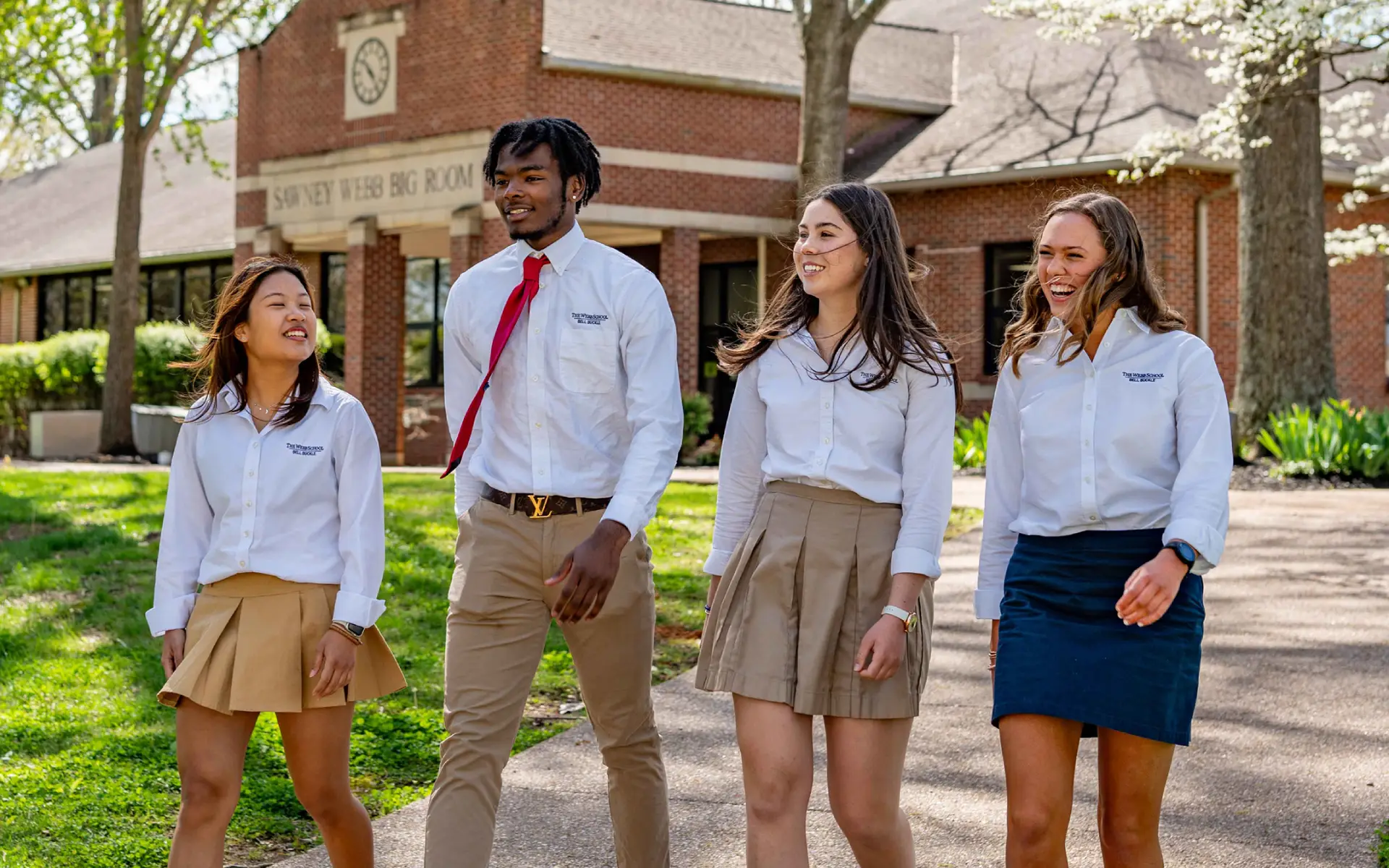 Four students wearing school uniforms walking and smiling on a sunny school campus with a brick building in the background.