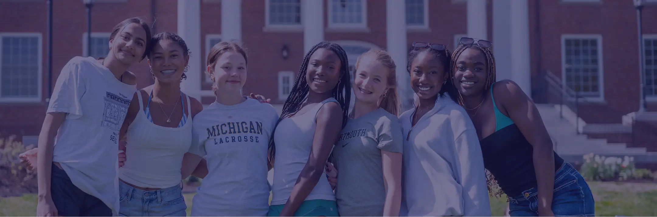 Group of seven diverse college-age women standing closely and smiling outdoors in front of a brick building with white columns.