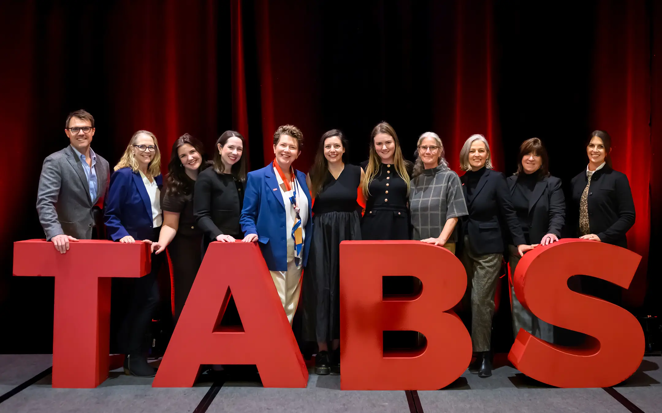 Group of eleven people standing behind large red letters spelling 'TABS' on a stage with red curtains.