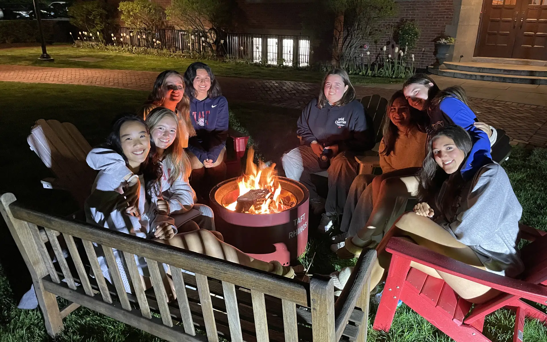 Group of eight young adults sitting around a glowing fire pit outdoors at night, smiling and enjoying the warmth.