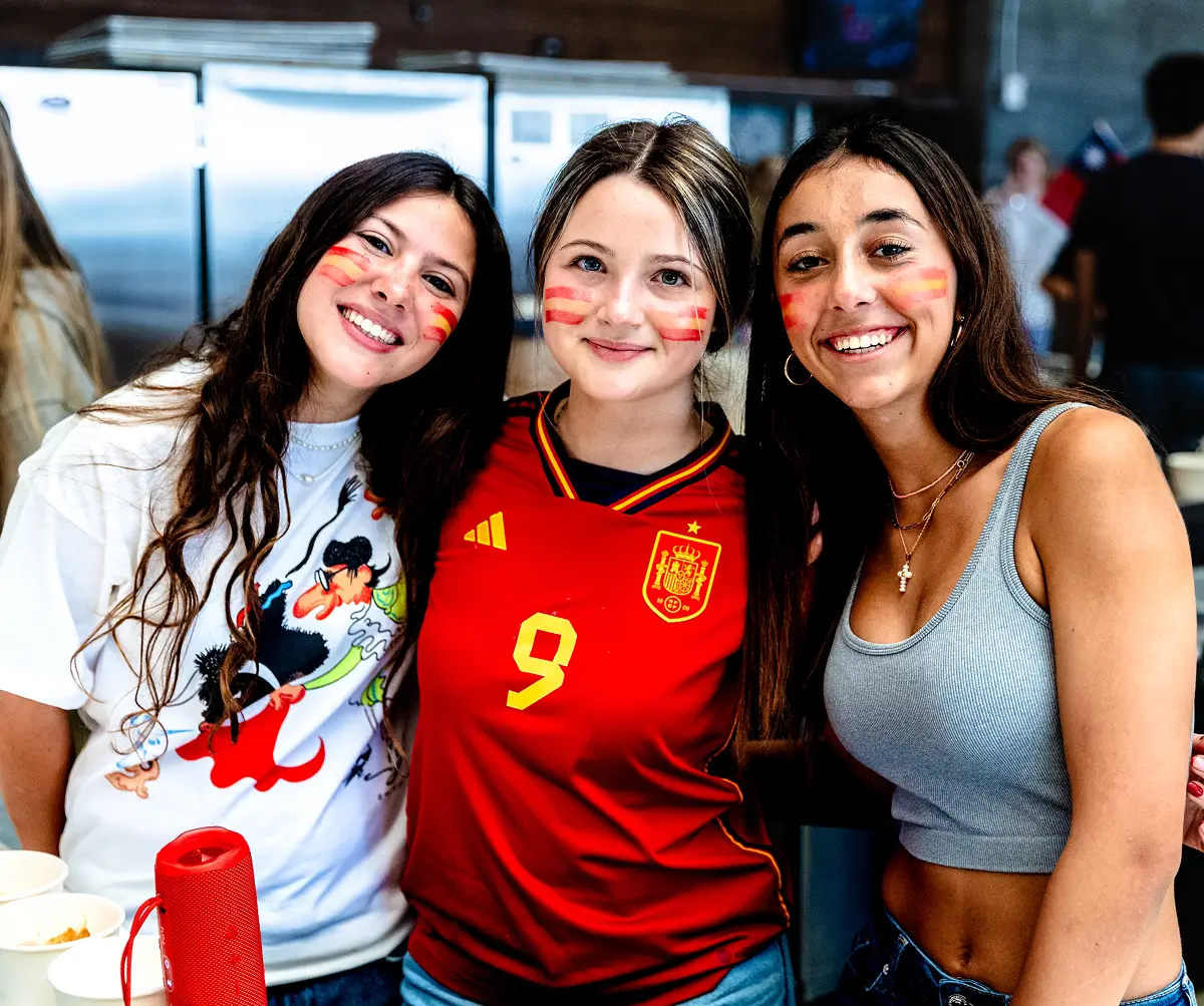 Three young women smiling with Spanish flag face paint, one wearing a red Spain soccer jersey.