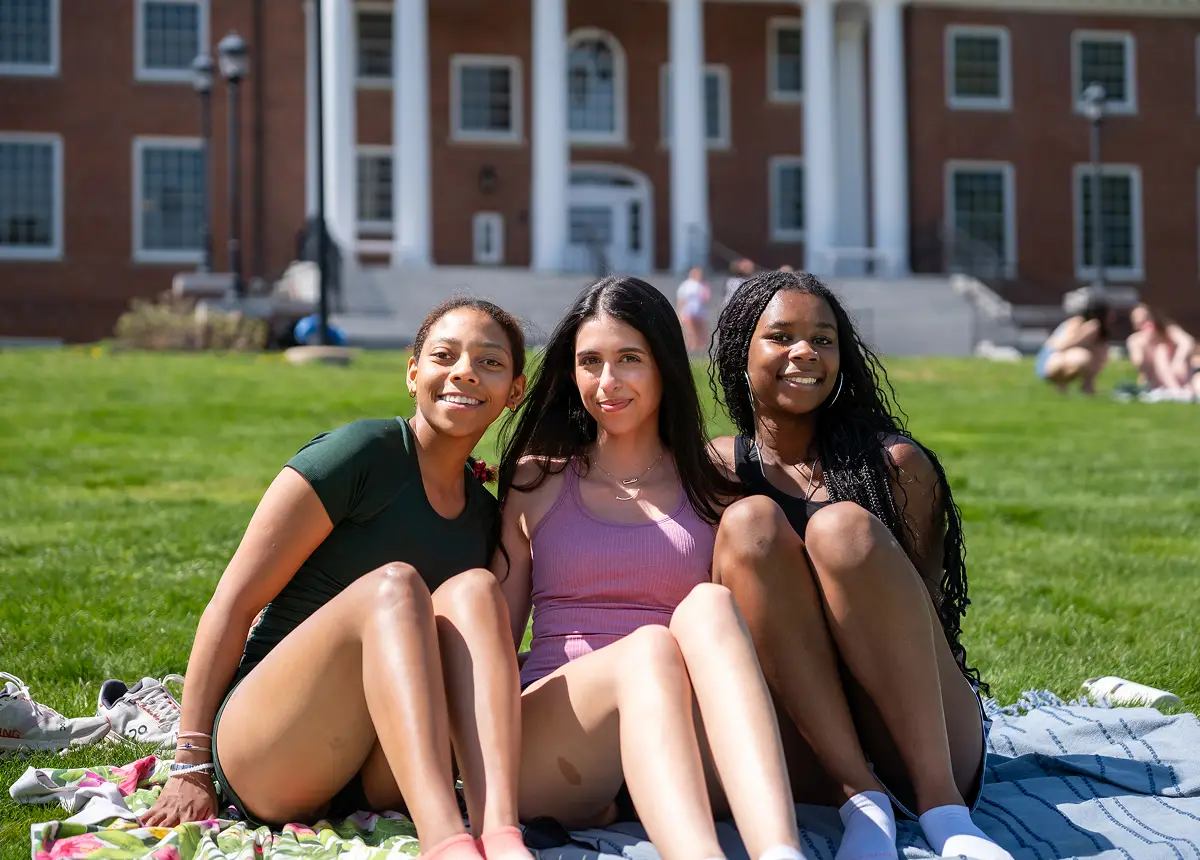 Three young women sitting on blankets on green grass in front of a brick building with white columns on a sunny day.