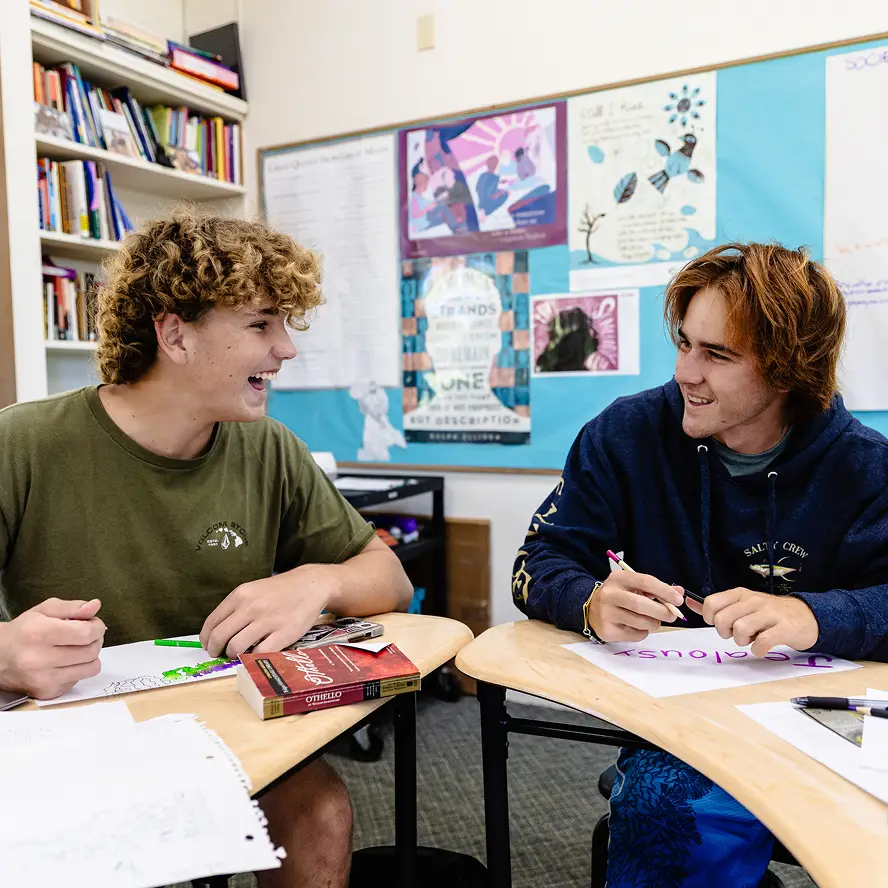 Two teenage boys smiling and talking while sitting at desks in a classroom with bookshelves and a bulletin board behind them.