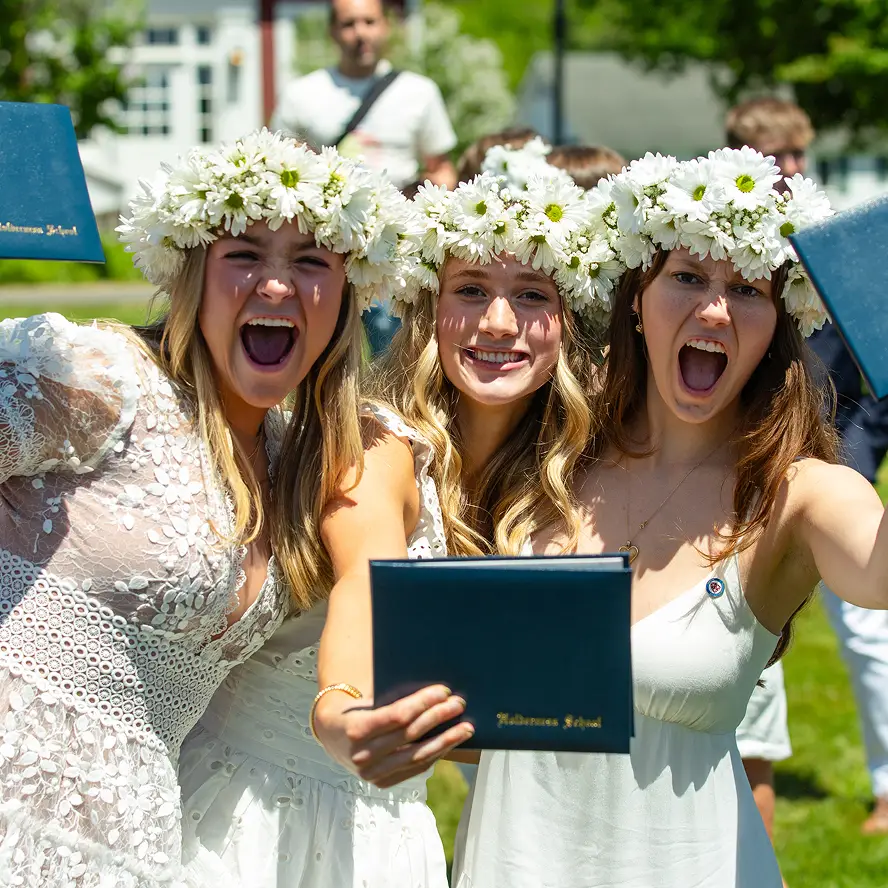 Three young women wearing white dresses and flower crowns holding diploma covers outdoors at a graduation celebration.